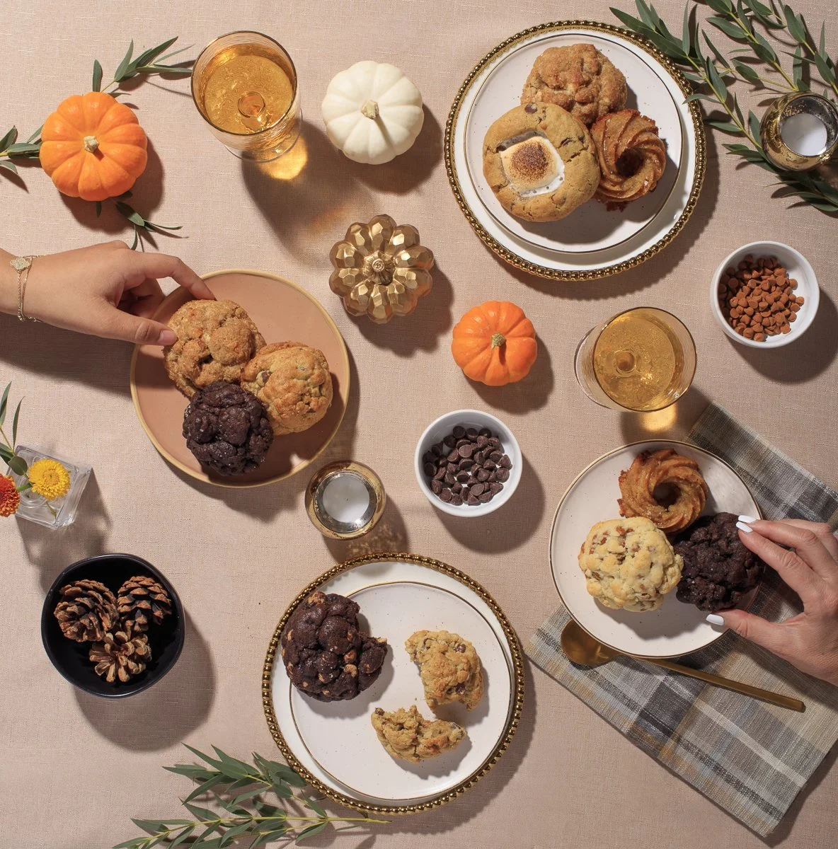 A table set with holiday-themed cookies, small pumpkins, pinecones, glasses of white wine, and bowls of chocolate chips and candy. Two people are reaching for cookies on plates.