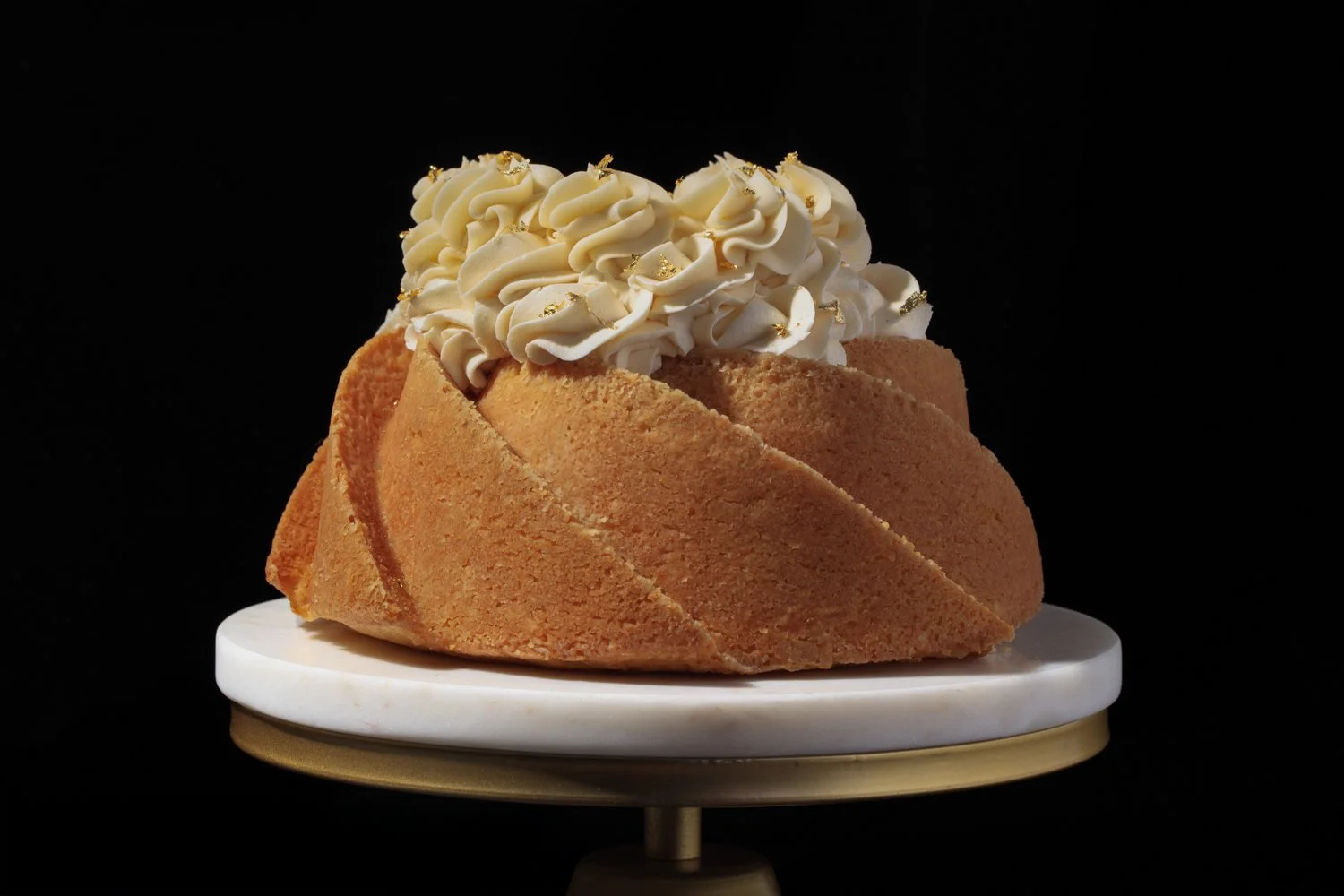 Cake with a sliced design, topped with white whipped cream ruffles and gold sprinkles, on a white and gold cake stand against a black background. Food stylist and photographer serving San Diego 