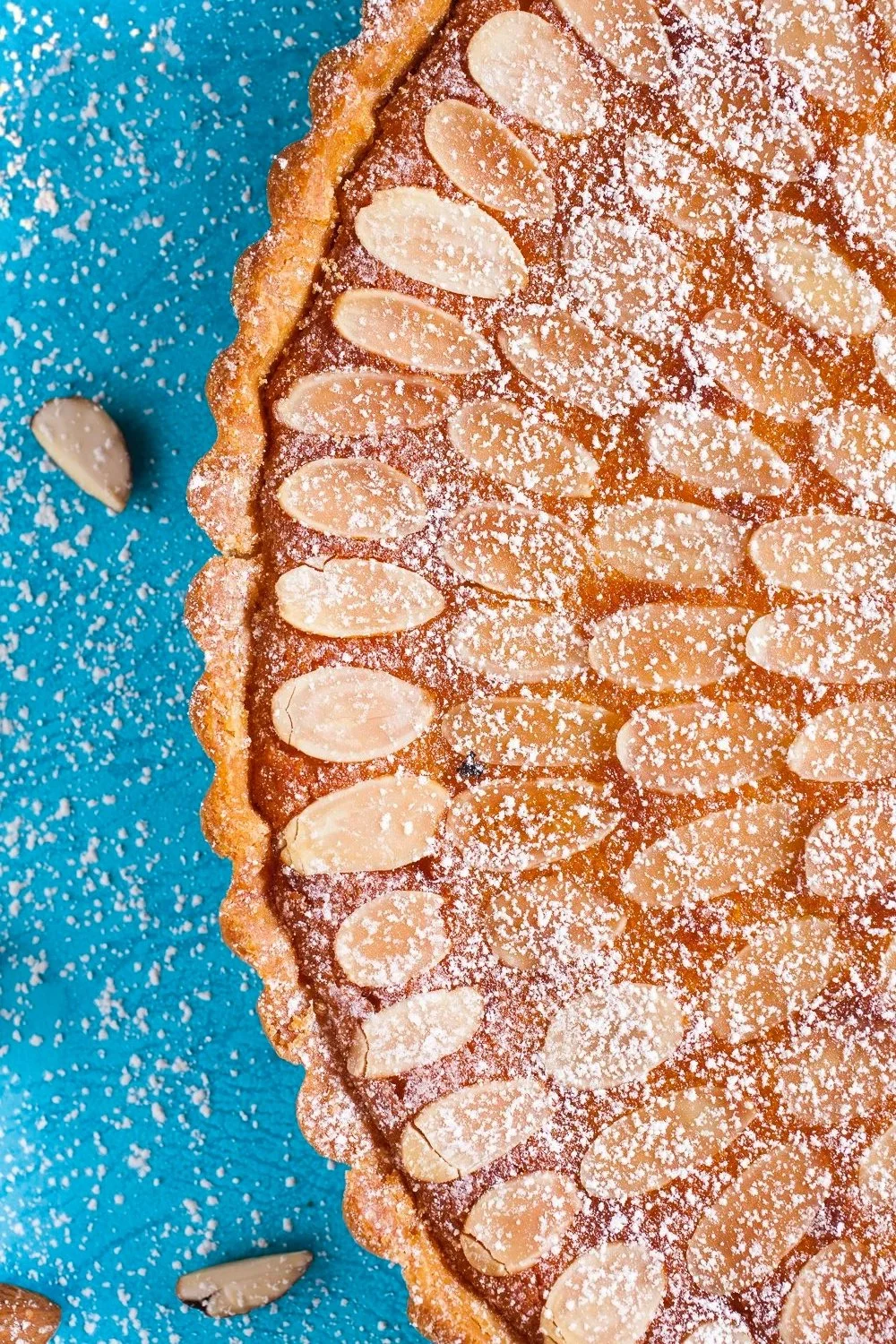Close-up of an almond tart on a blue surface, dusted with powdered sugar, with scattered almond slices around. Orange County food and lifestyle photographer 