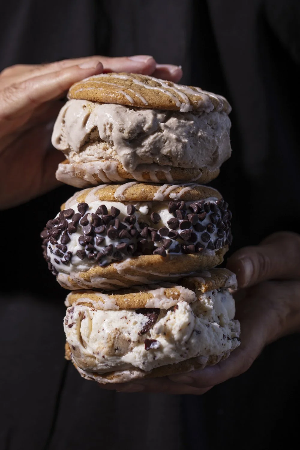 A person holding a stack of four assorted cookies with ice cream and chocolate chips, topped with white icing.