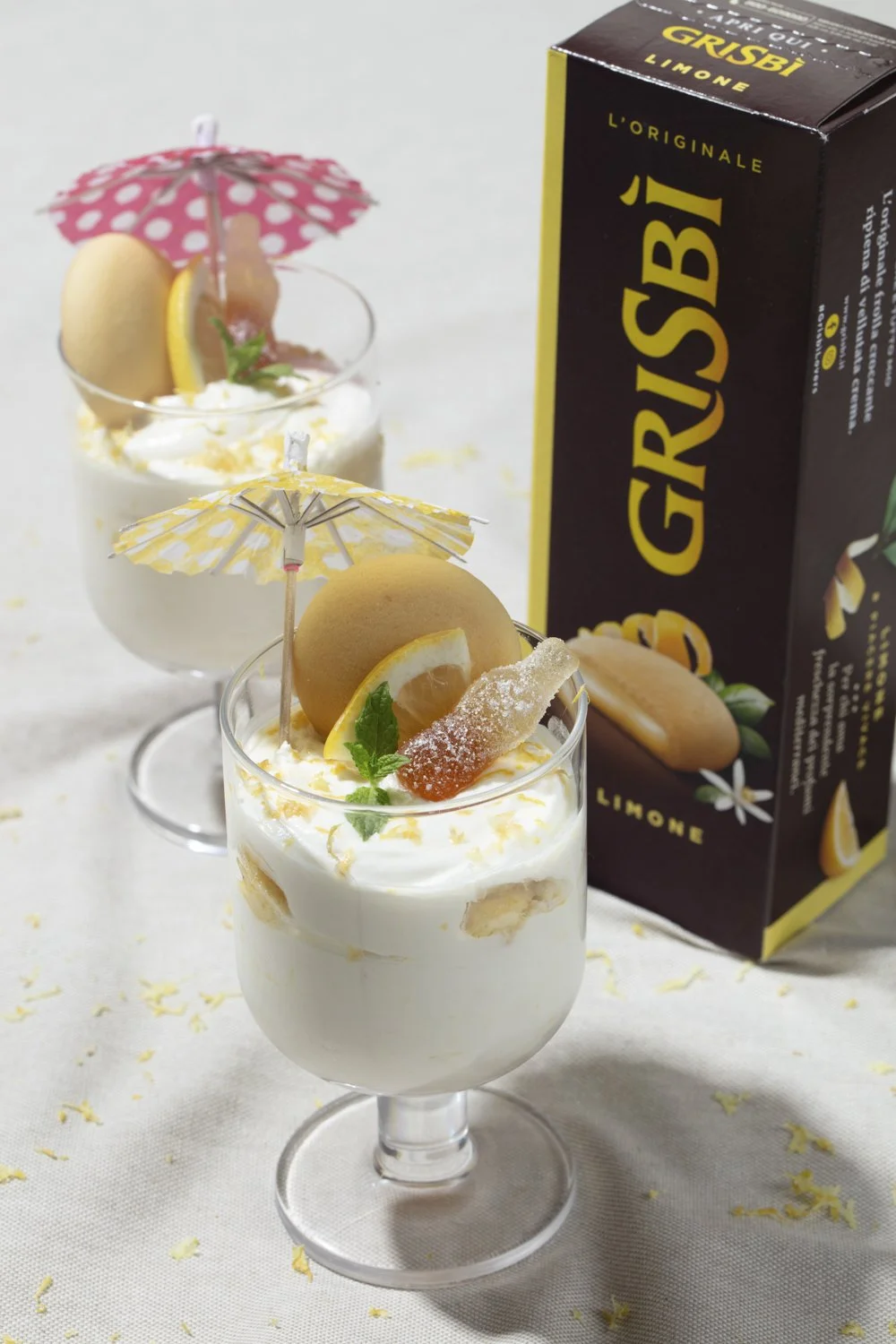 Two dessert glasses containing creamy lemon mousse topped with lemon slices, cookies, mint leaves, and small decorative umbrellas, with a carton of Grisbi lemon cookies in the background. Los Angeles food photographer and stylist 