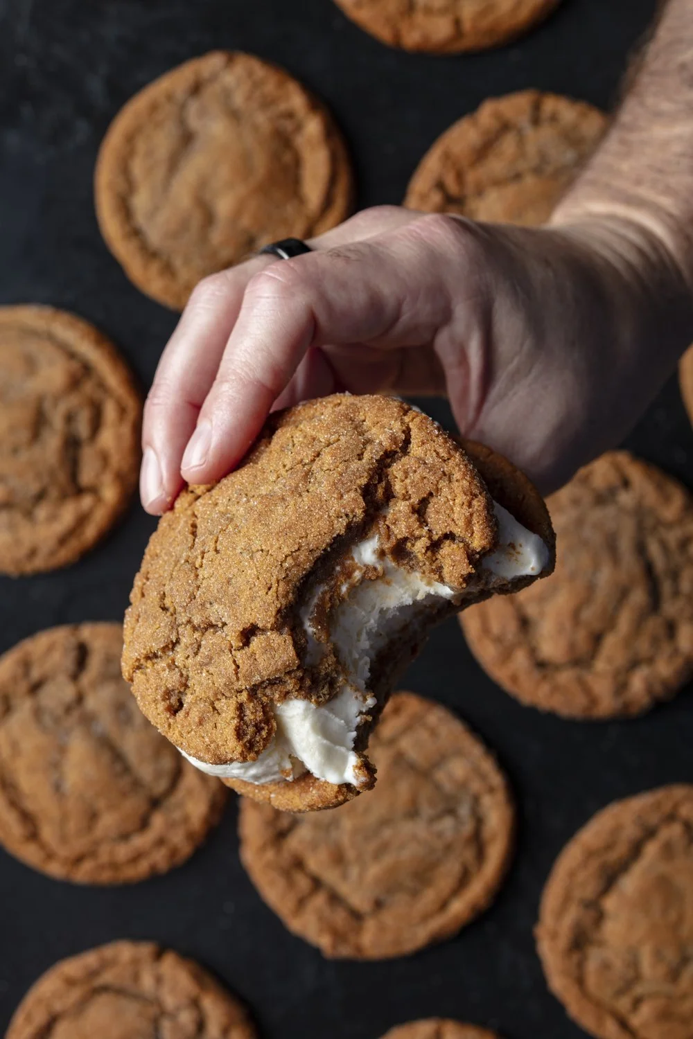 A hand holding a partially eaten chocolate chip cookie with melted ice cream inside, with more cookies on a dark surface in the background.