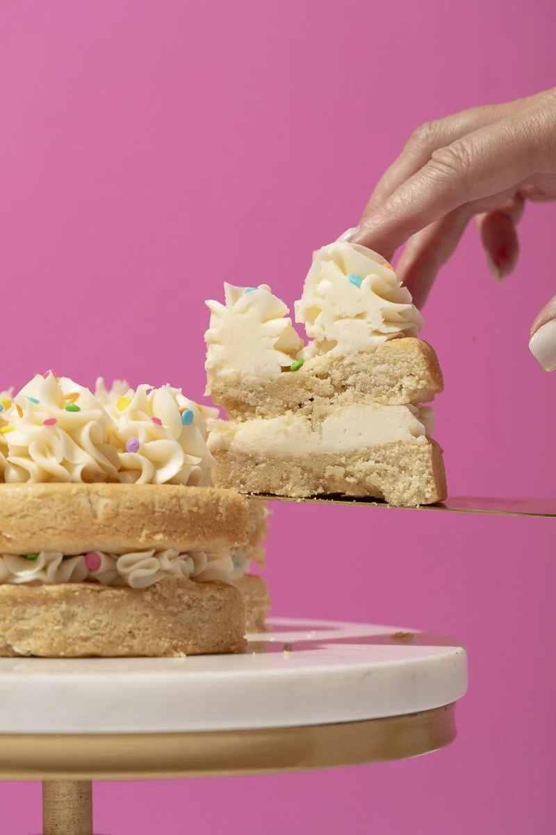 A hand is lifting a slice of cake with vanilla frosting and colorful sprinkles from a layered cake on a white cake stand against a pink background.  San Diego