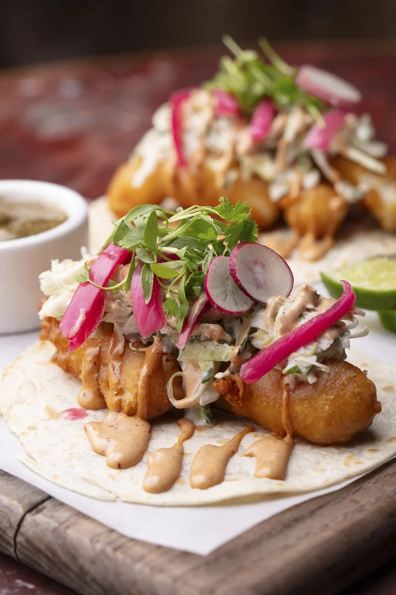 Tacos with fried fish, shredded lettuce, sliced radishes, pickled red onions, cilantro, and drizzled creamy sauce on a soft tortilla, served with lime wedges.
