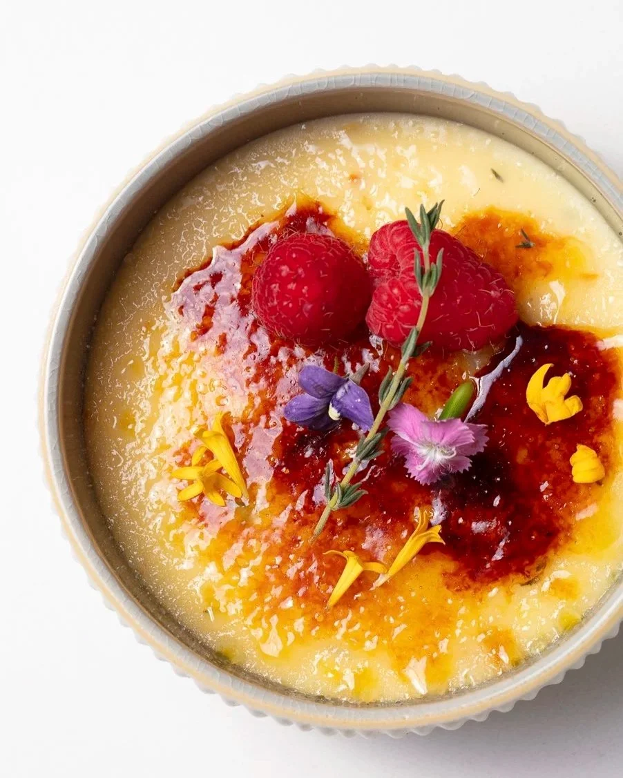 A dessert with a yellow custard base topped with raspberries, edible flowers, and a drizzle of red sauce.