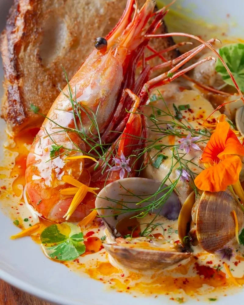 Seafood dish with shrimp, clams, and fish, garnished with herbs and edible flowers, served with a slice of toasted bread.