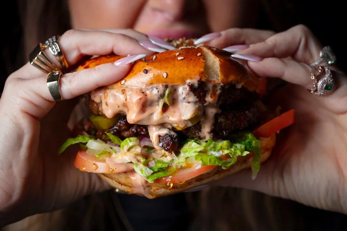 Person holding and about to eat a large burger with lettuce, tomato, cheese, a beef patty, and sauce in a bun with sesame seeds.