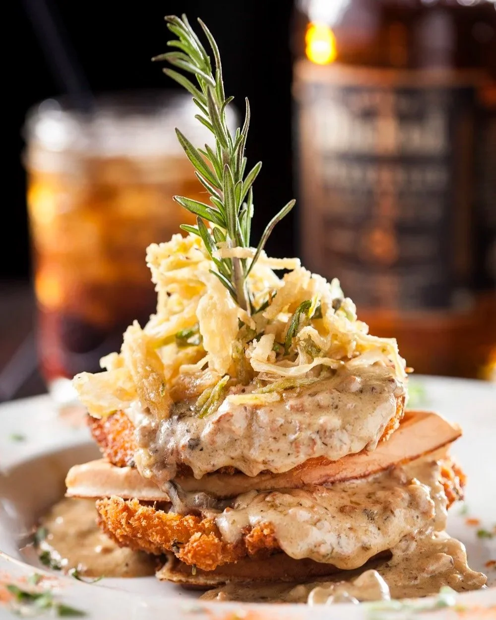 A plate of breaded fried chicken topped with gravy, mashed potatoes, and shredded cabbage, garnished with a sprig of rosemary.