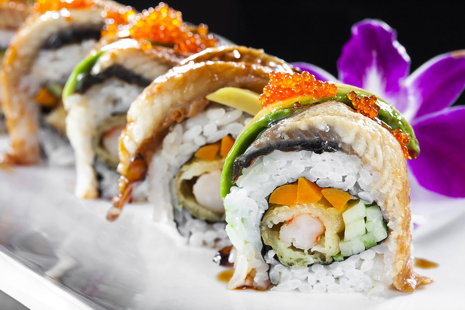 Close-up of a colorful sushi roll topped with fish, avocado, and orange fish roe, with a purple flower in the background.