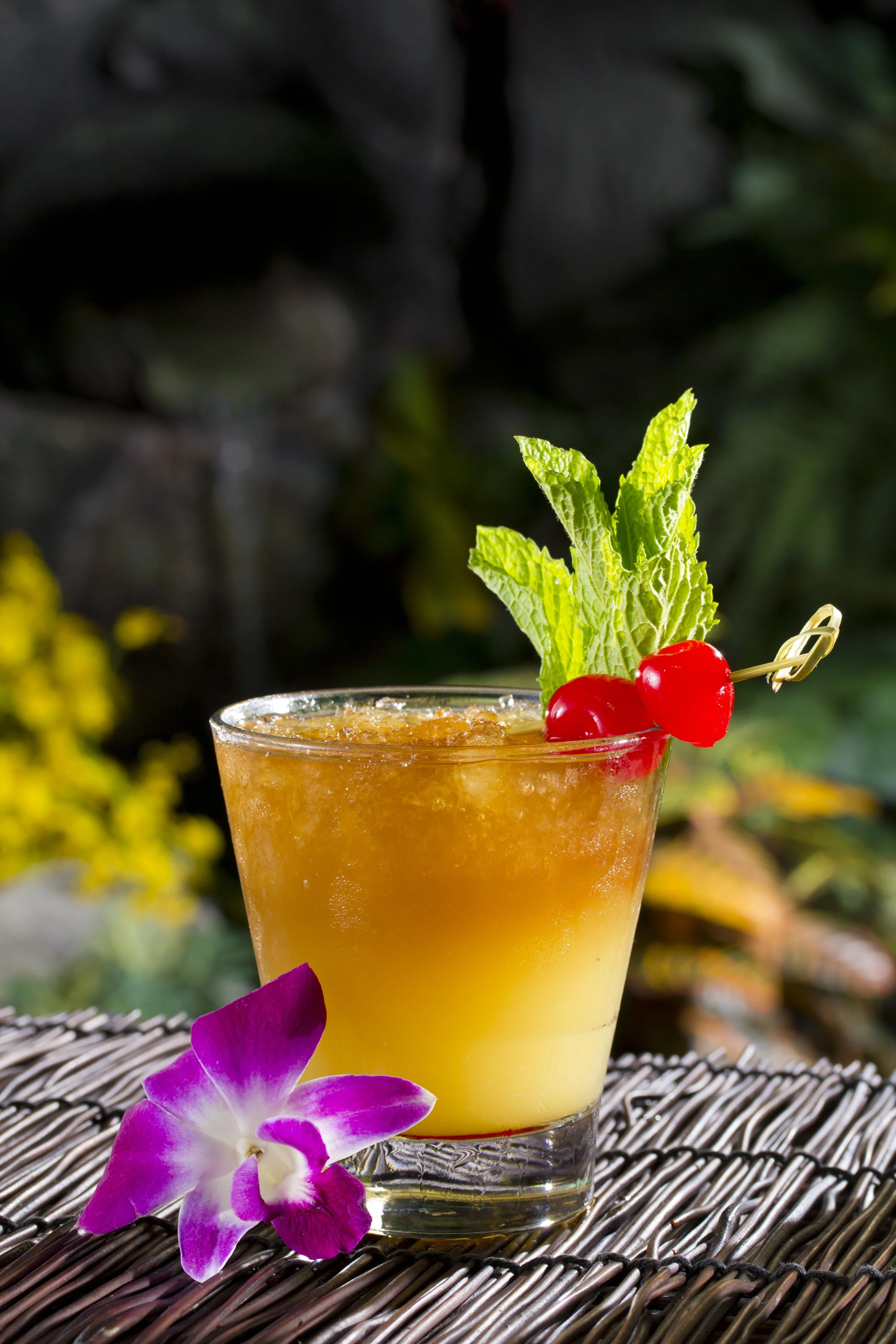 A colorful cocktail drink garnished with mint leaves and red cherries, with purple and white orchid flowers beside it, set on a woven surface with a blurred outdoor background.