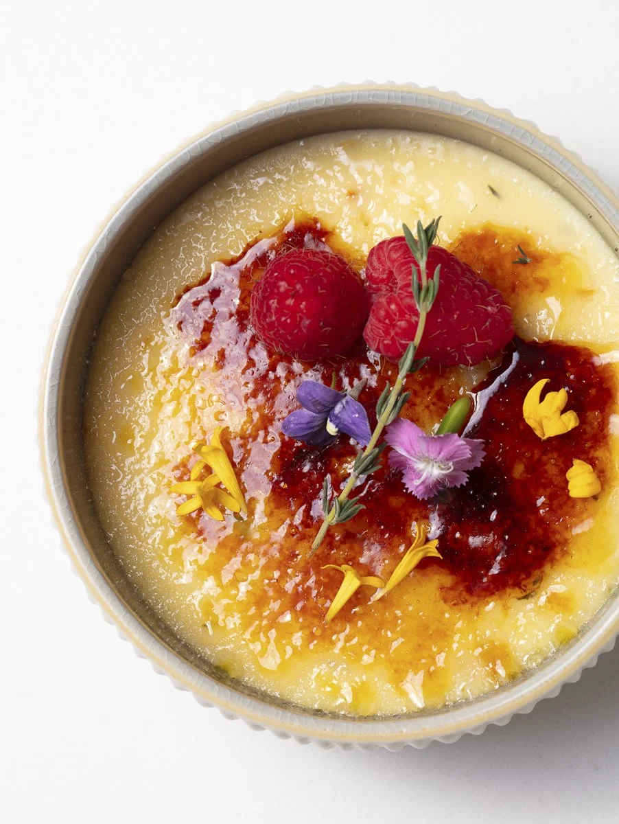 A dessert with a smooth, creamy surface topped with raspberries, edible flowers, and a drizzle of red sauce.