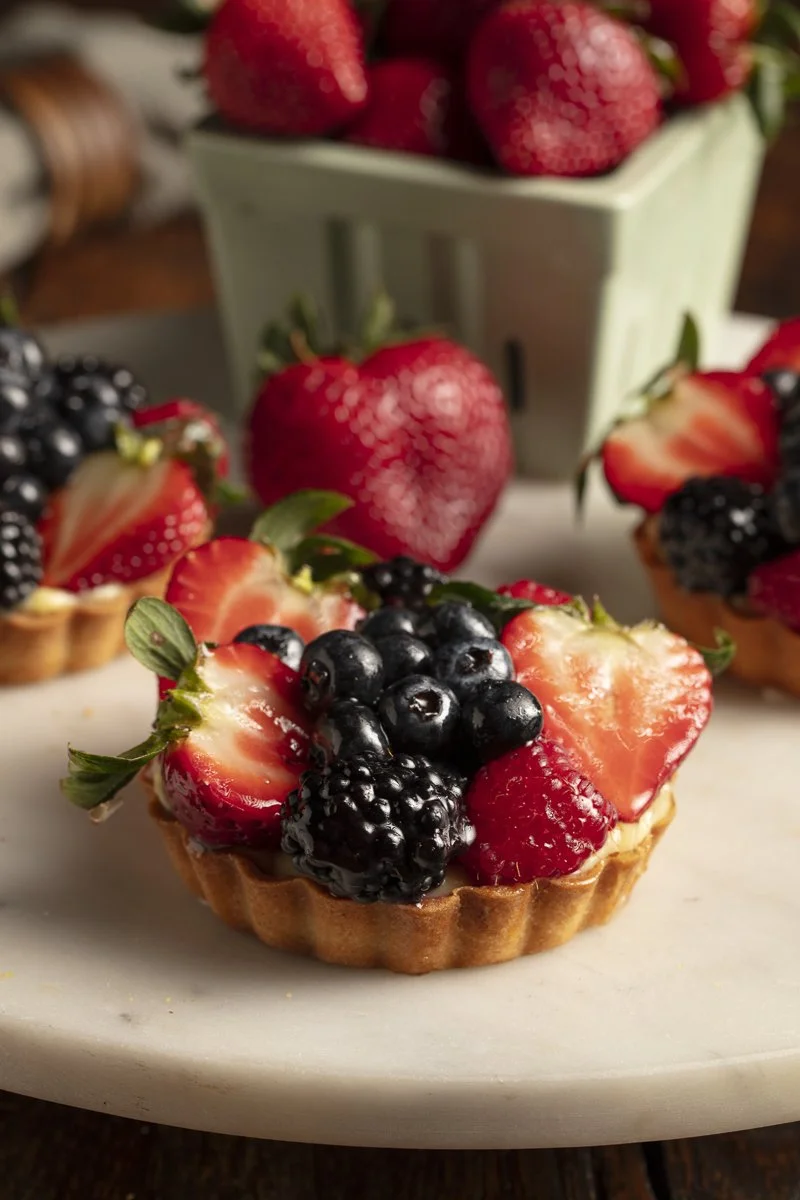 A fruit tart topped with sliced strawberries, blackberries, blueberries, and raspberries on a white plate. Scatch bakery - Orange County food photographer and stylist 