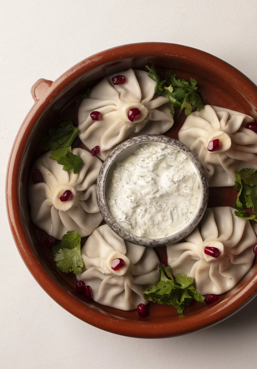 A round terracotta dish containing six khinkali dumplings garnished with pomegranate seeds and cilantro, served with a side of creamy herb sauce.