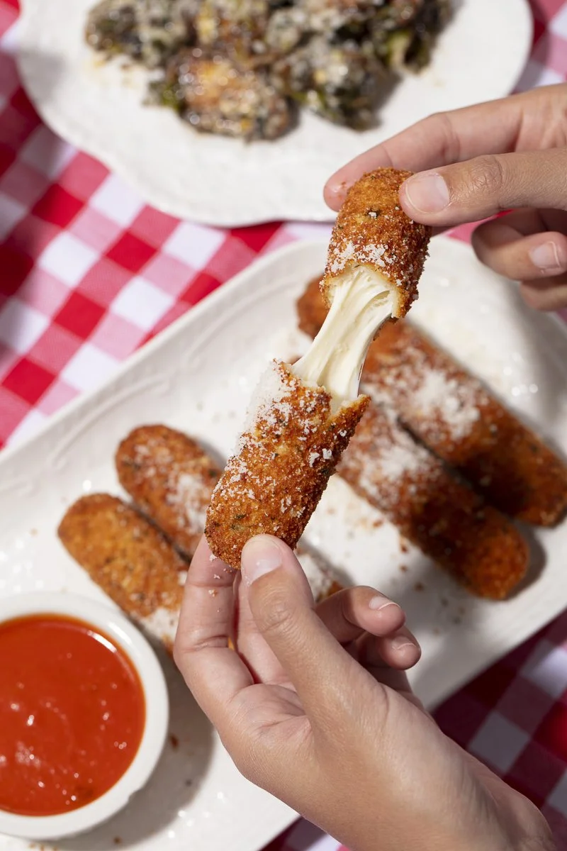 Person holding a fried cheese stick with melted cheese stretching from it, on a white plate with several more cheese sticks and a small dish of red dipping sauce, all on a red and white checkered tablecloth.