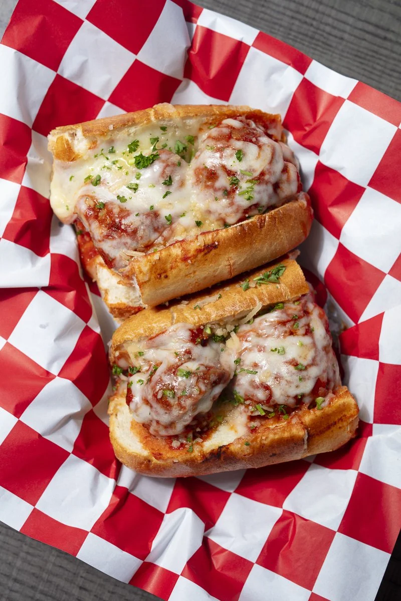 Cheeseburger sub sandwich with melted cheese and meatballs, garnished with chopped herbs, on a checkered paper-lined tray.