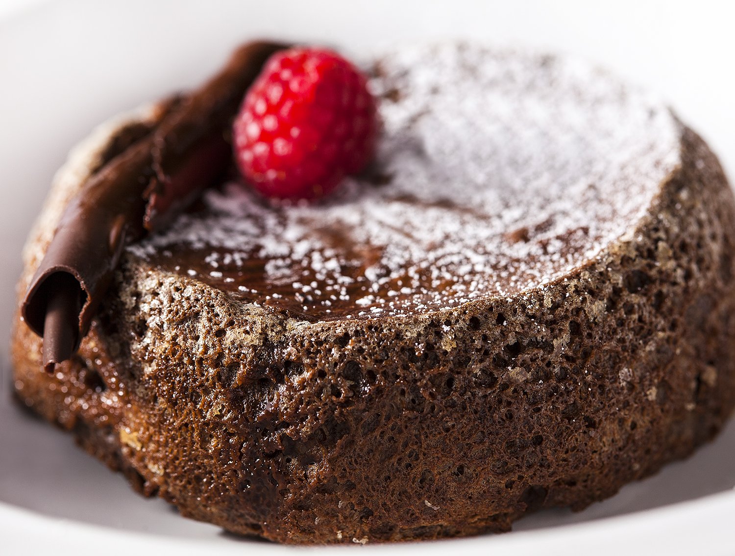Close-up of a chocolate lava cake topped with a strawberry, dusted with powdered sugar, with a chocolate curl on the side.