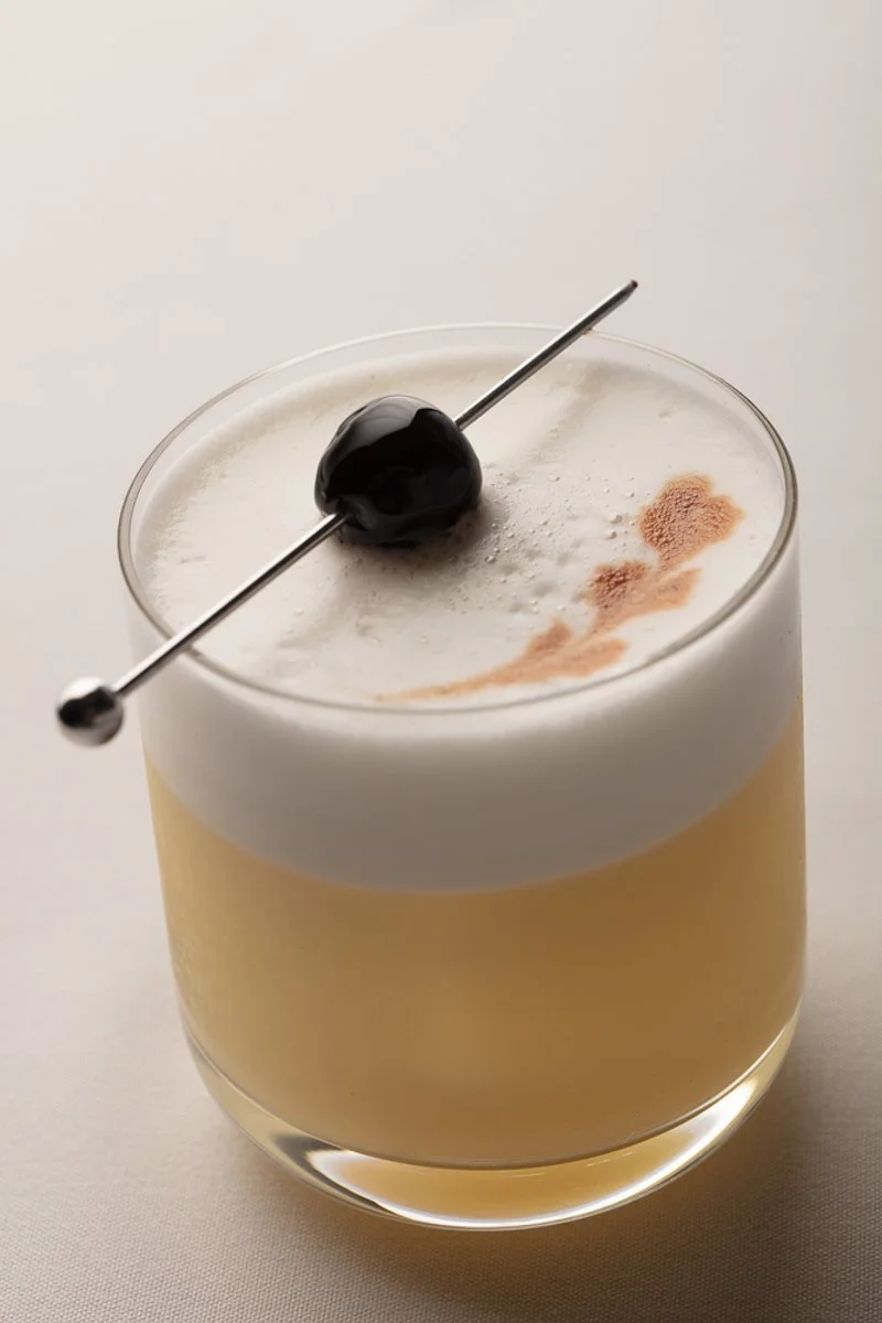 A layered cocktail in a clear glass with a frothy white top, garnished with a black cherry on a metal skewer and a sprinkle of cinnamon or spices on top.