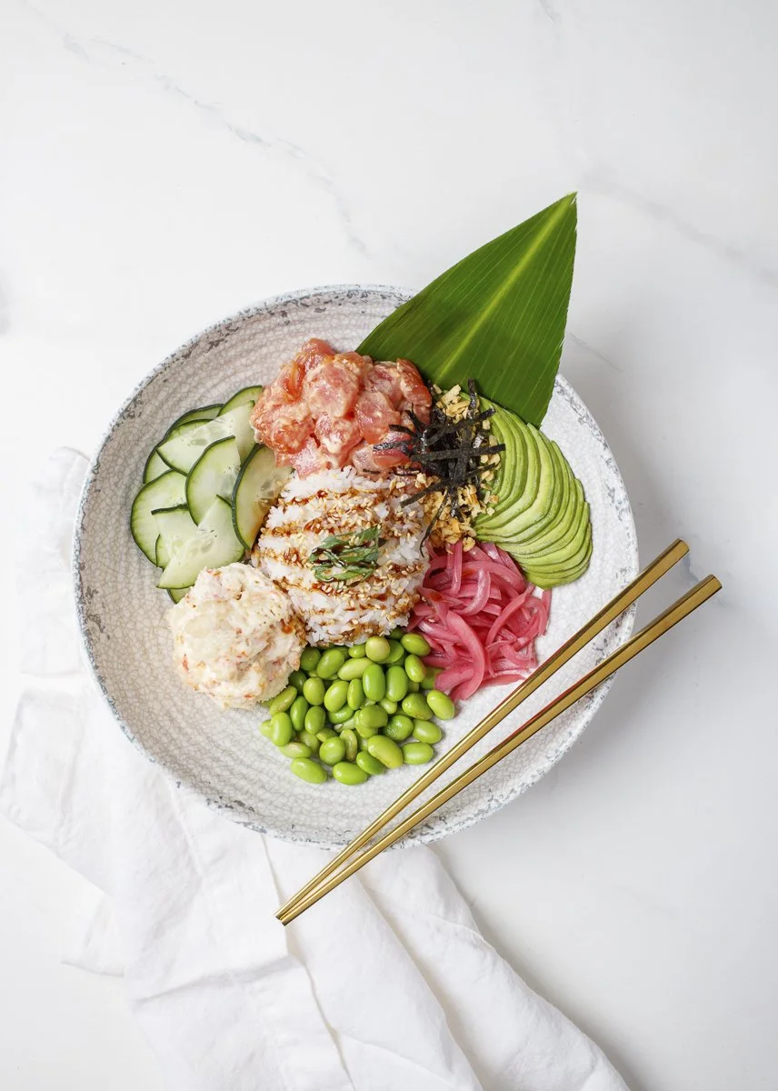 A bowl of assorted sushi and sashimi with cucumber slices, avocado, pickled ginger, wasabi, shredded nori, and green edamame beans, with a green leaf and chopsticks on a white background.