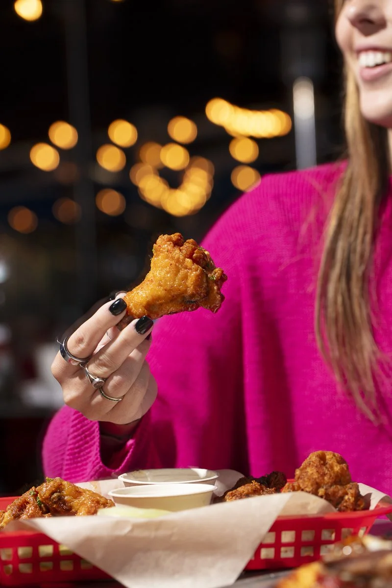 Person in a pink sweater holding a piece of fried chicken with a smile, in a restaurant or dining setting with blurred lights in the background.