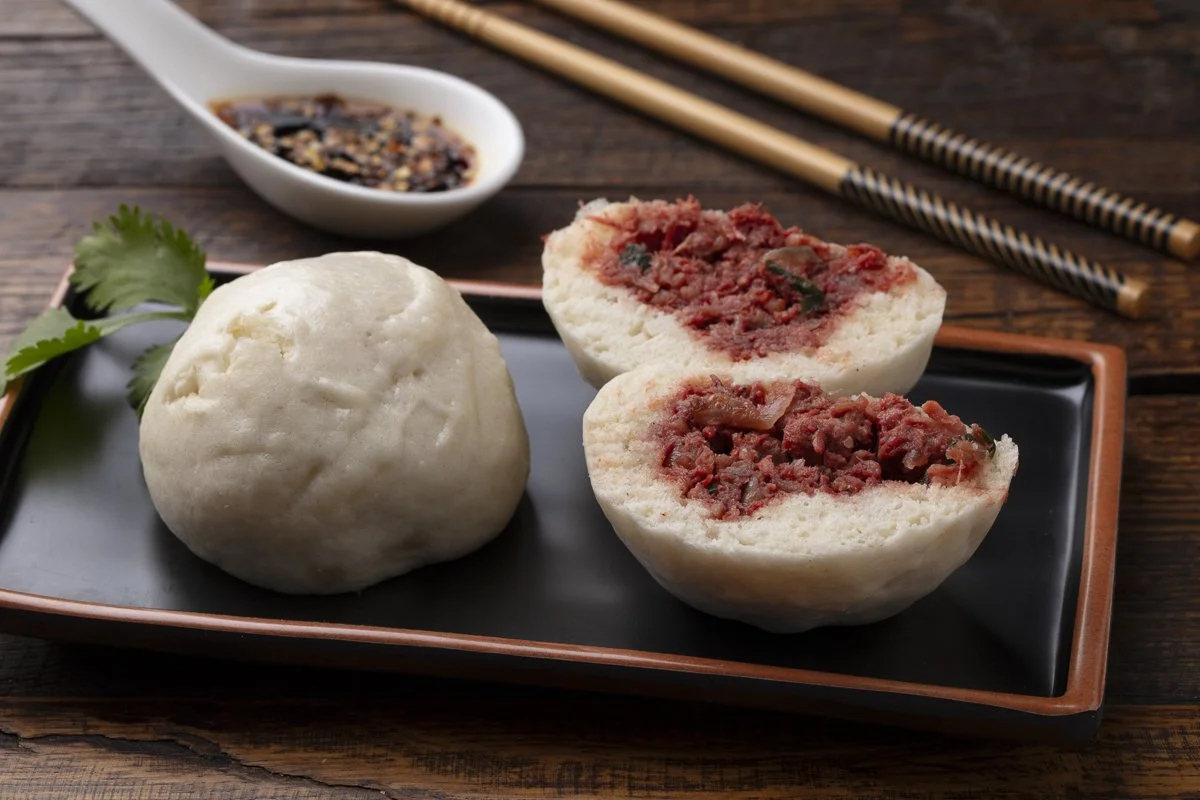 Two steamed Chinese pork buns, one whole and one cut in half to reveal the meat filling, on a rectangular black and brown plate. A small dish of soy sauce with sesame seeds and chopped chilies, with a pair of chopsticks on a wooden table.