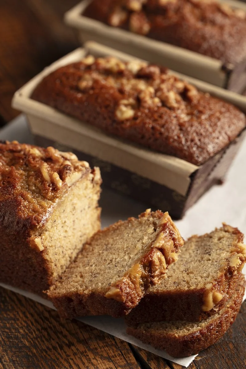 Close-up of a sliced banana nut bread with walnuts on top, placed on parchment paper on a wooden surface, with a whole loaf in the background. Scratch Bakery - Food photographer and stylist - Plate to PIxel 