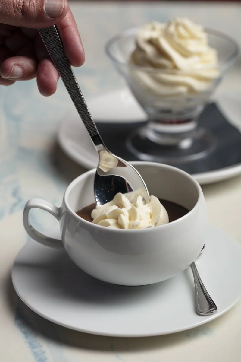A person holding a spoon with whipped cream above a white mug of hot chocolate topped with whipped cream, with another bowl of whipped cream in the background.