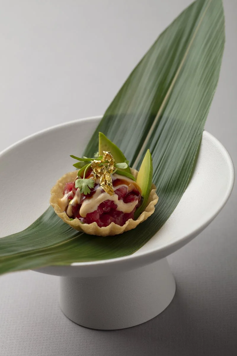 A small tart with red fruit filling, topped with cream or sauce, garnished with herbs and gold leaf, placed on a large green leaf in a white bowl.