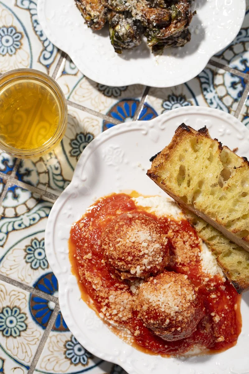A plate with two Italian meatballs covered in marinara sauce and sprinkled with grated cheese, along with slices of garlic bread, and a glass of white wine on a patterned table.