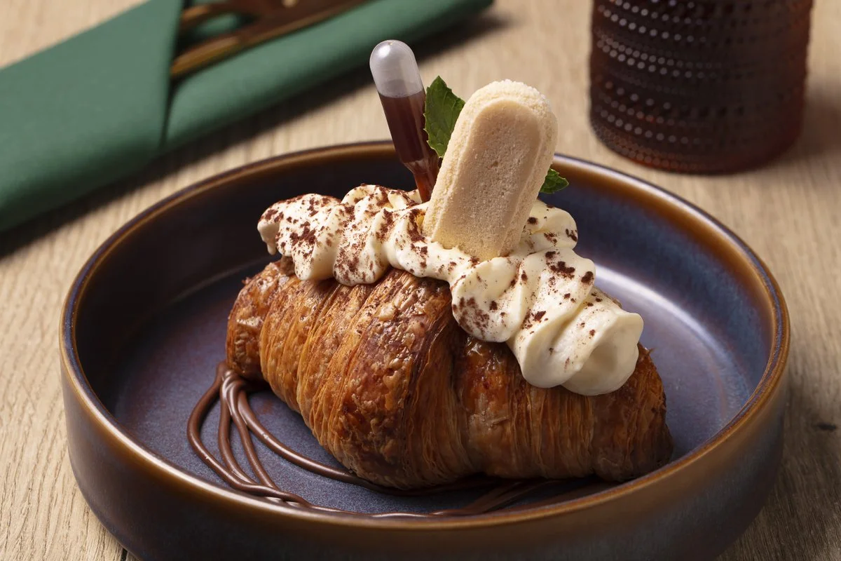 A croissant dessert topped with whipped cream, cocoa powder, a cookies and cream ice cream sandwich, a chocolate capsule, a mint leaf, and chocolate drizzle on a blue ceramic plate.