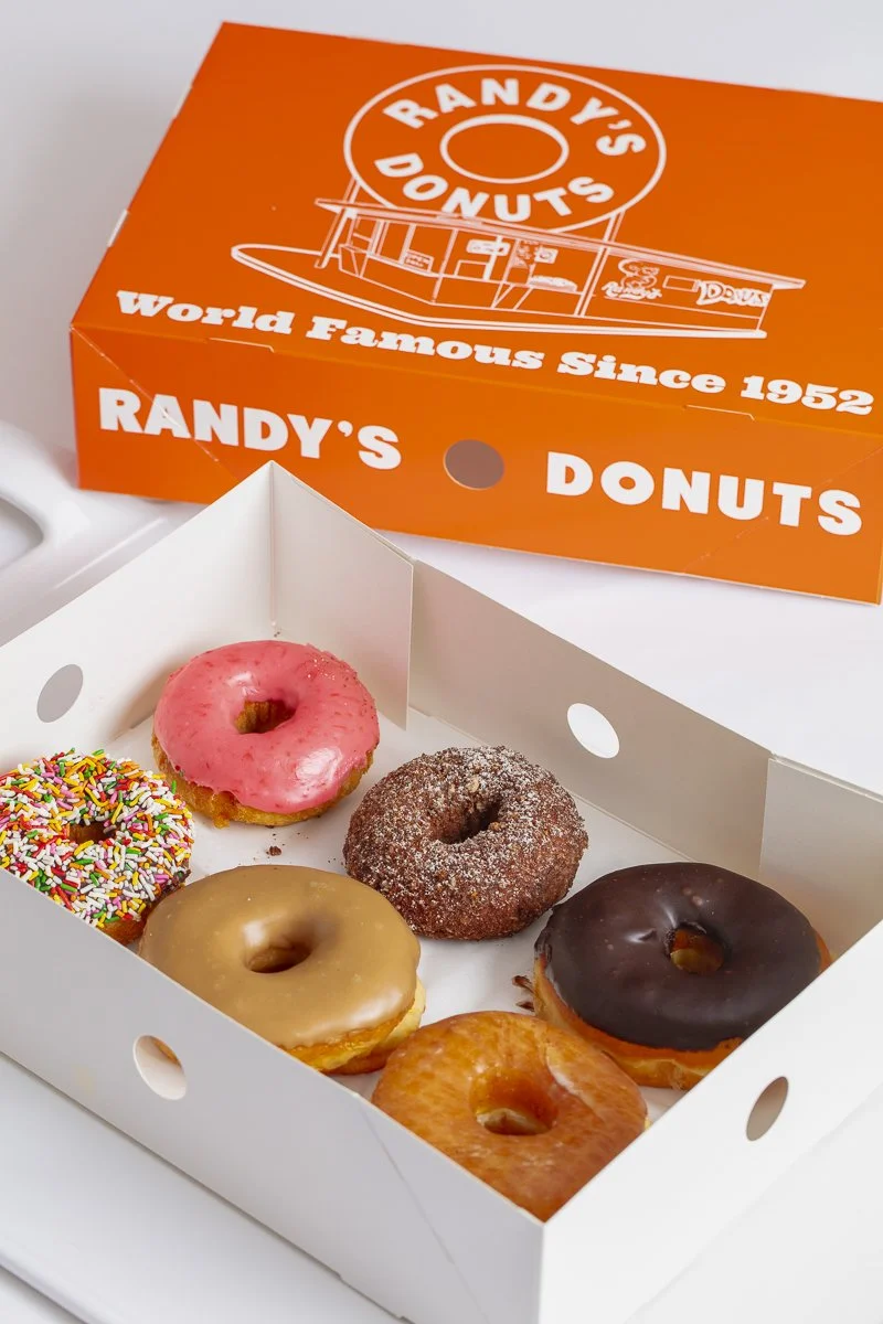 A white box containing six assorted donuts with pink glaze, sprinkles, sugar coating, chocolate glaze, caramel glaze, and plain glazed, placed next to an orange Randy's Donuts box with logo and text. Los Angeles food and product photographer and styl