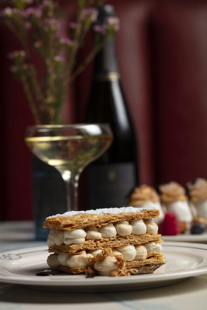 A plate with layered cream-filled pastries, a glass of white wine, a wine bottle, and a plate of desserts in the background with a red wall and flowers.