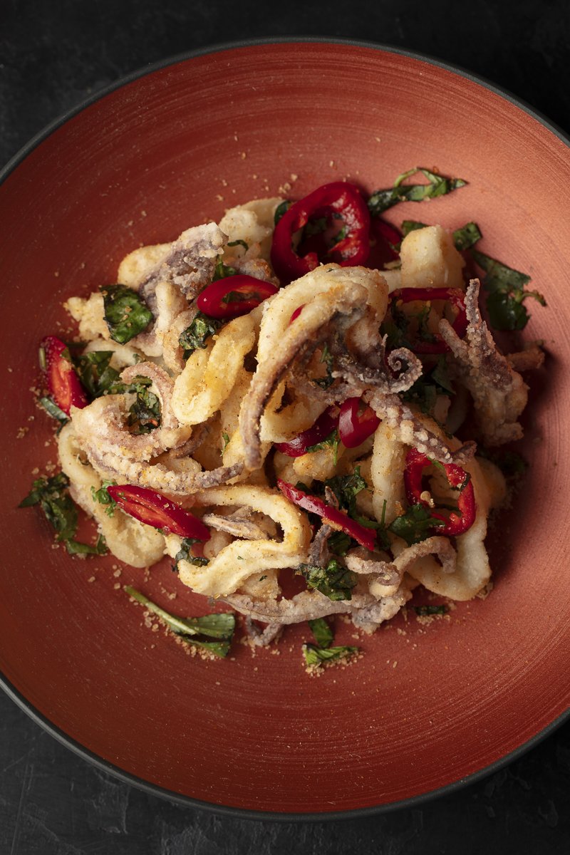 Fried calamari garnished with sliced red chili peppers and chopped green herbs, served in a red bowl.