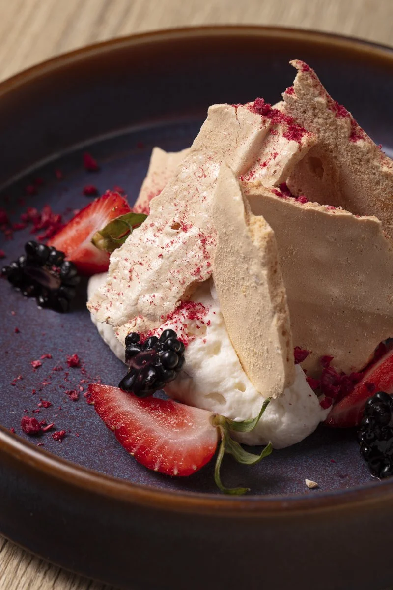 Close-up of a dessert with meringue, whipped cream, strawberries, blackberries, and red crumbs on a dark plate. Food photographer and stylist pastry photo shoot in Orange County 