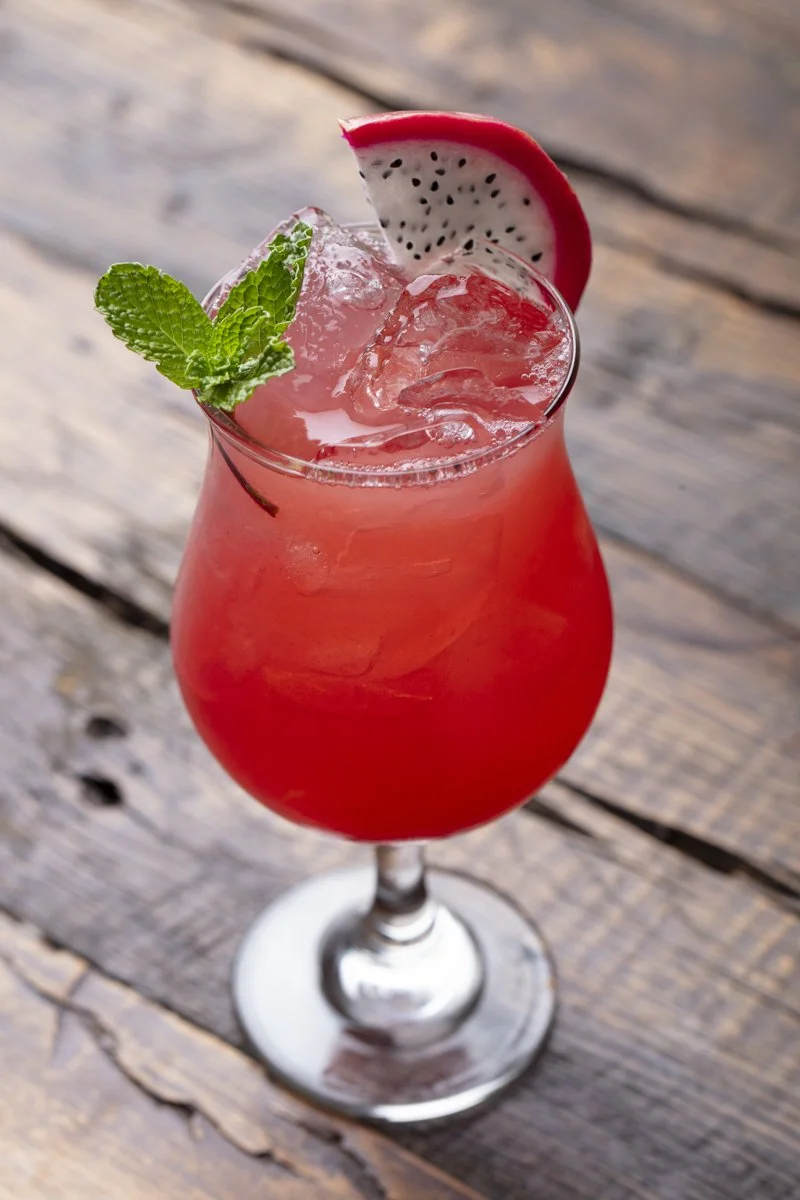 A pink cocktail garnished with a slice of dragon fruit and a sprig of mint, served over ice in a stemmed glass on a wooden surface.