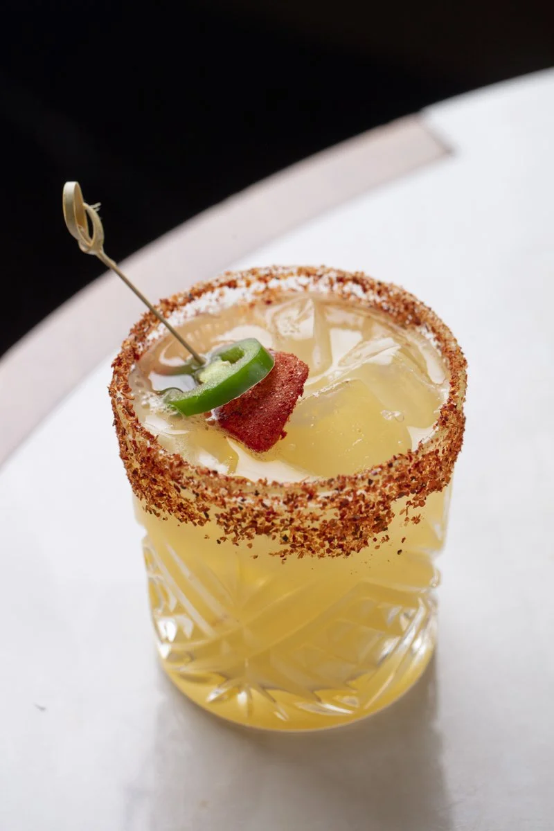 A yellow cocktail in a faceted glass with a salted rim, garnished with a jalapeño slice and a small square of bacon, served on a white surface.