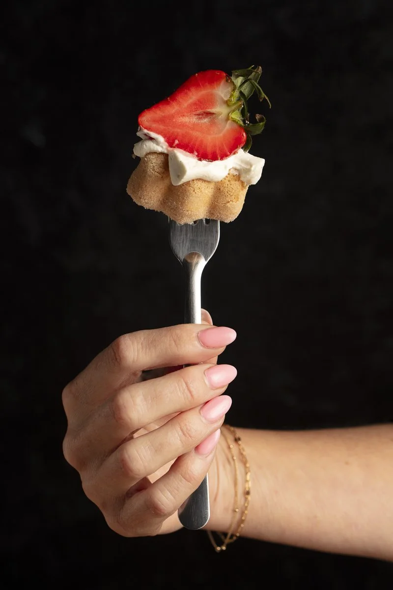 A fork held by a hand with pink nails, with a bite-sized piece of cake topped with whipped cream, a strawberry half, and a small green leaf against a black background. Batch and Box photo shoot by food photographer and stylist Plate to Pixel 