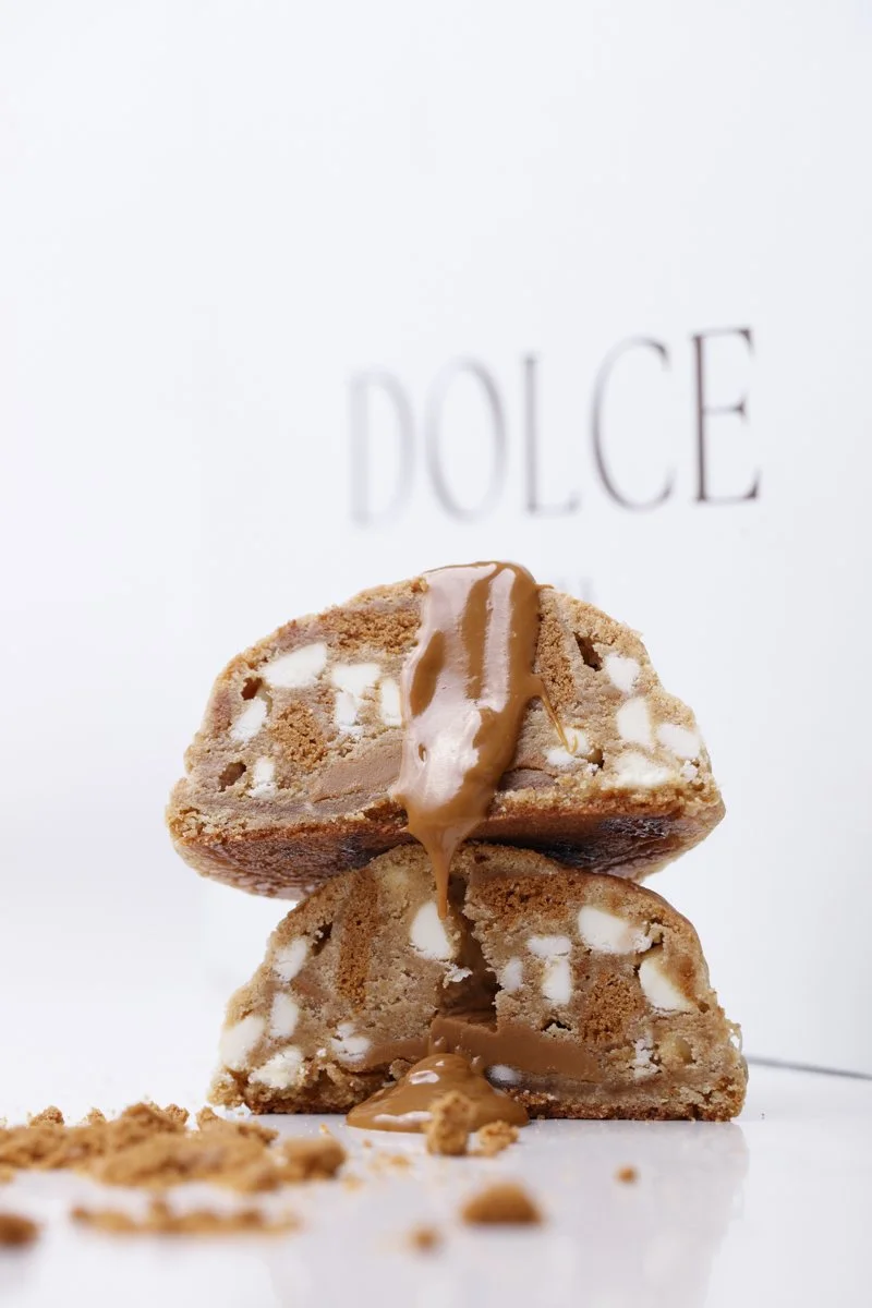 Stacked cookie pieces with caramel and white icing, with a white background and blurred text that reads "DOLCE."