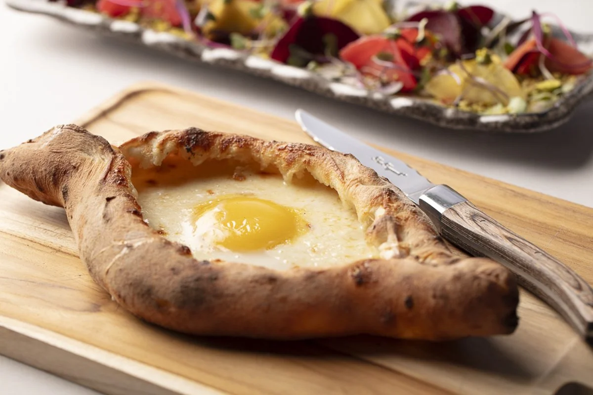 Khachapuri with a runny egg yolk in the center on a wooden board, with a knife nearby, and a tray of salad in the background.