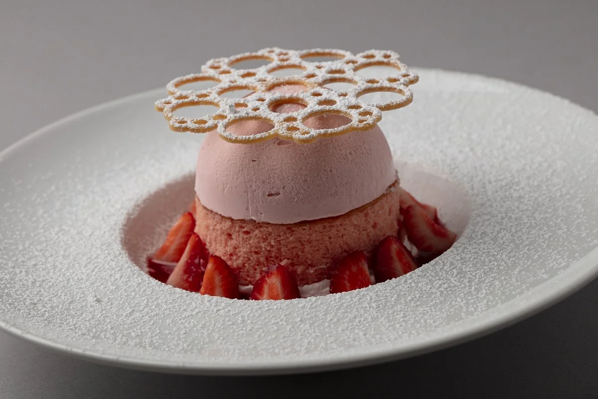 A pink strawberry mousse dessert with sliced strawberries at the base, topped with a decorative sugar lace on a white plate dusted with powdered sugar.