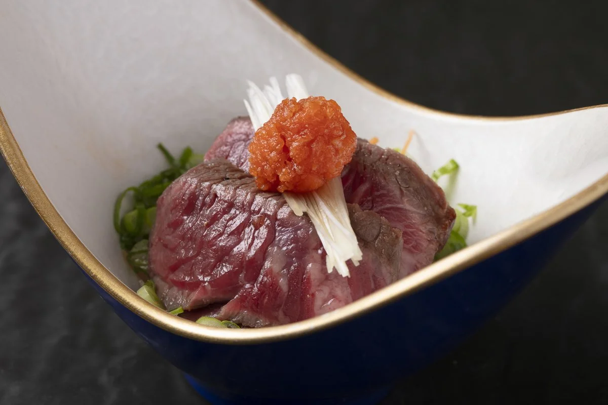 Sliced raw beef topped with a dollop of orange roe, garnished with white shredded garnish and chopped green onions in a ceramic bowl