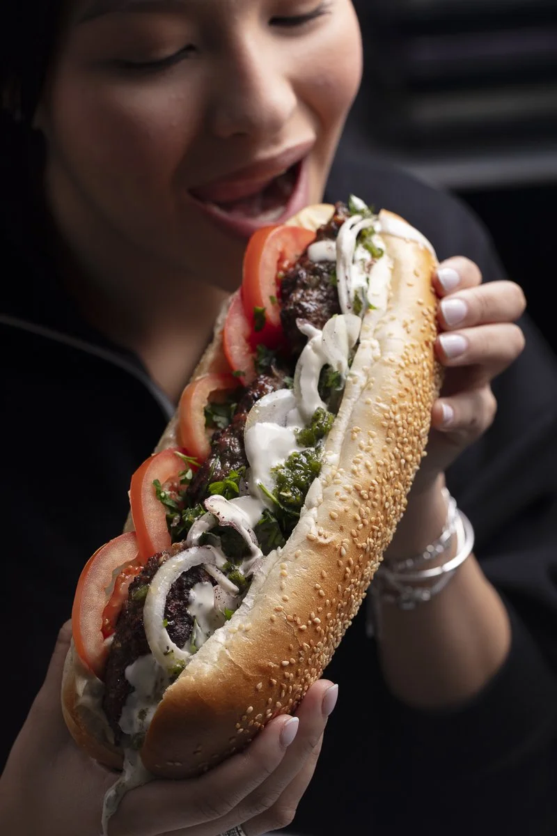 Person holding and about to eat a large sandwich with tomatoes, onions, lettuce, cheese, and bread.