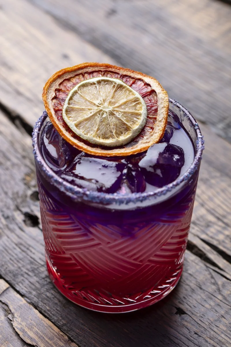 A colorful drink in a textured glass garnished with a dried lemon slice and a dried orange slice, placed on a rustic wooden surface.