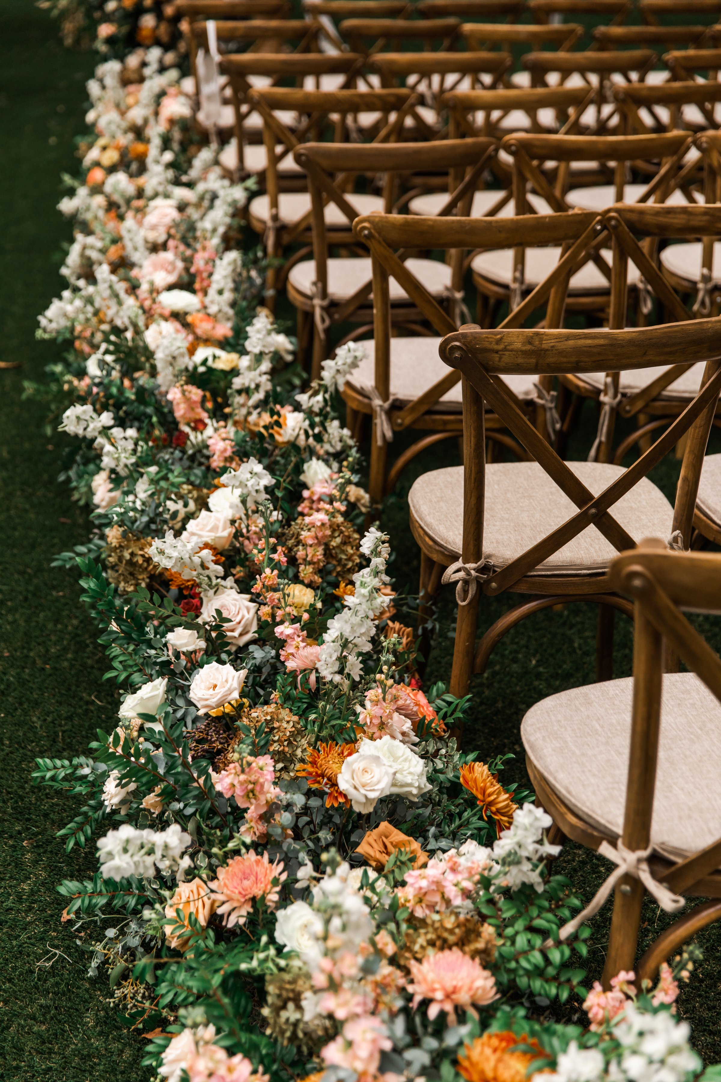 Lush floral hedges create aisle meadows for this fall wedding. Florals feature hues of terra cotta, blush, yellow, copper, and neutrals. Beautiful garden roses, dahlias, and fall florals highlight these arrangements. Design by Rosemary and Finch in N