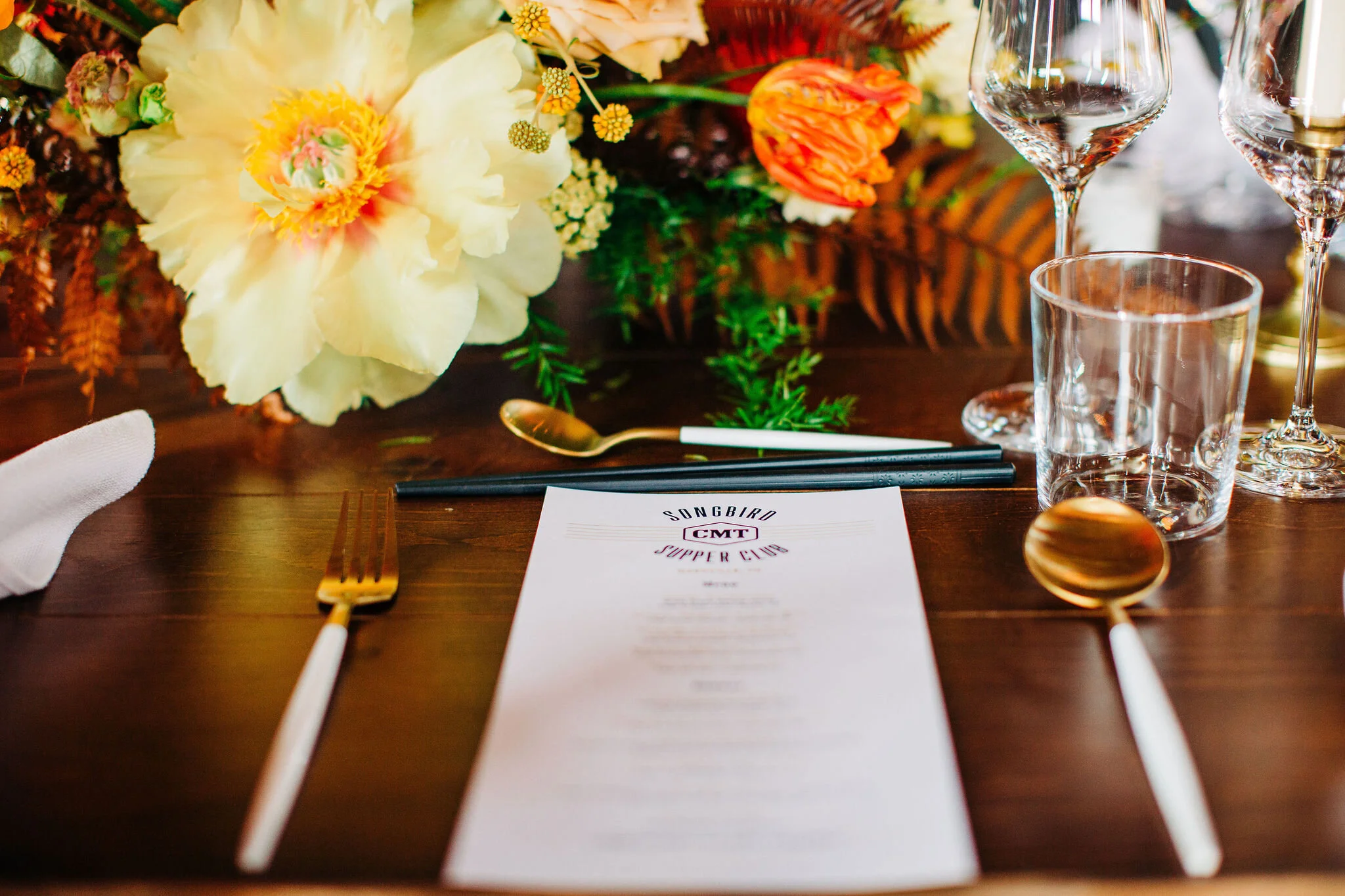 Yellow peonies, orange ranunculus, and golden fern floral centerpieces with brass candlesticks. Nashville wedding florist.