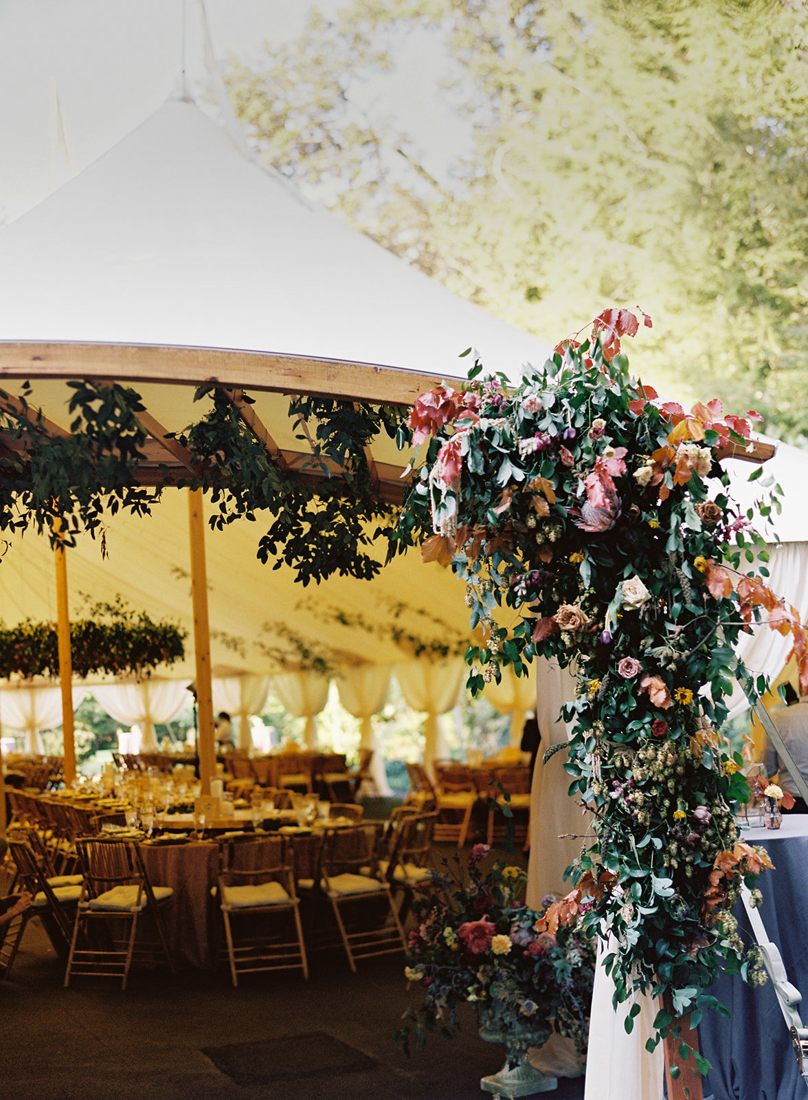 Tent entrance flower installation with lush vines, hops, fruiting pomegranate branches, greenery, wildflowers in shades of pink, mauve, copper, golden yellow, and soft blue. RT Lodge wedding floral designer.