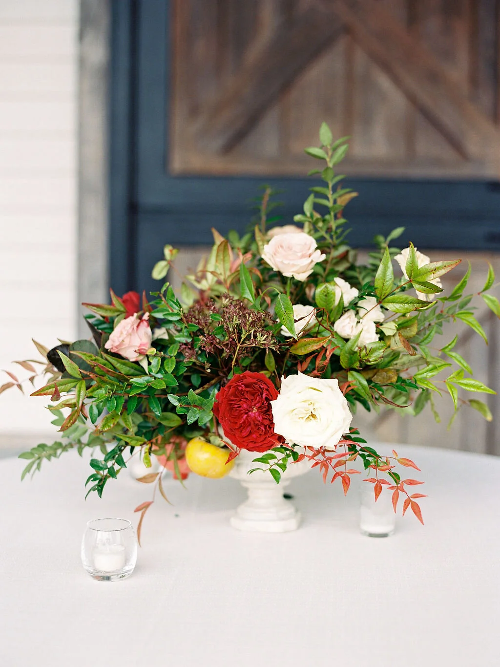 Low European garden urn overflowing with a natural floral arrangement of garden roses, ranunculus, festival bush, fruiting persimmon branches, organic greenery, and fall berries. Centerpiece for a Trinity View Farm wedding by Nashville florist, Rose…