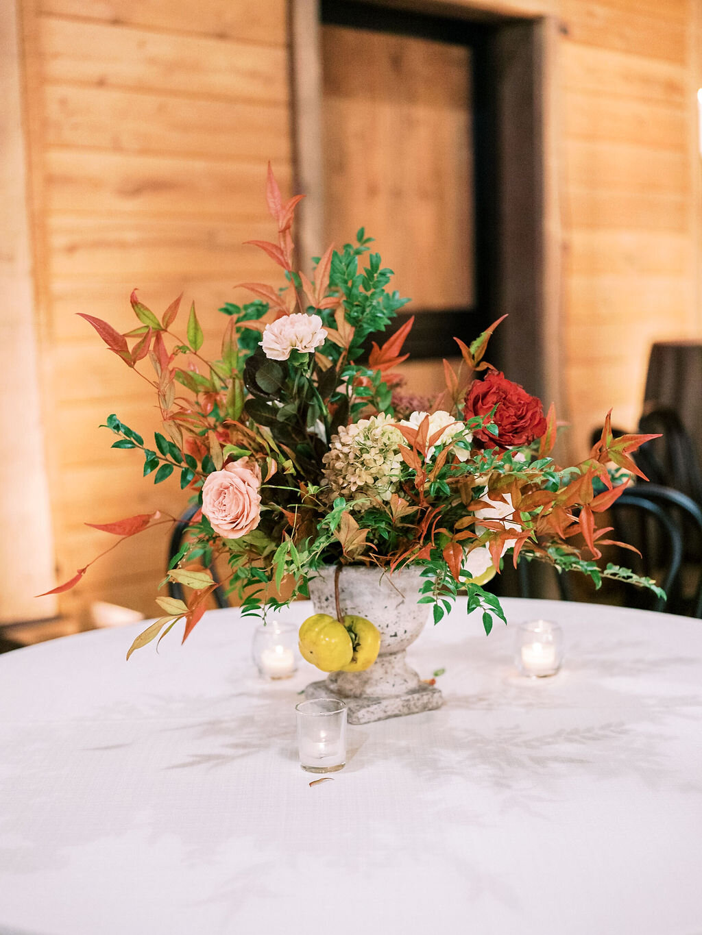 Low European garden urn overflowing with a natural floral arrangement of garden roses, ranunculus, festival bush, fruiting persimmon branches, organic greenery, and fall berries. Centerpiece for a Trinity View Farm wedding by Nashville florist, Rose…