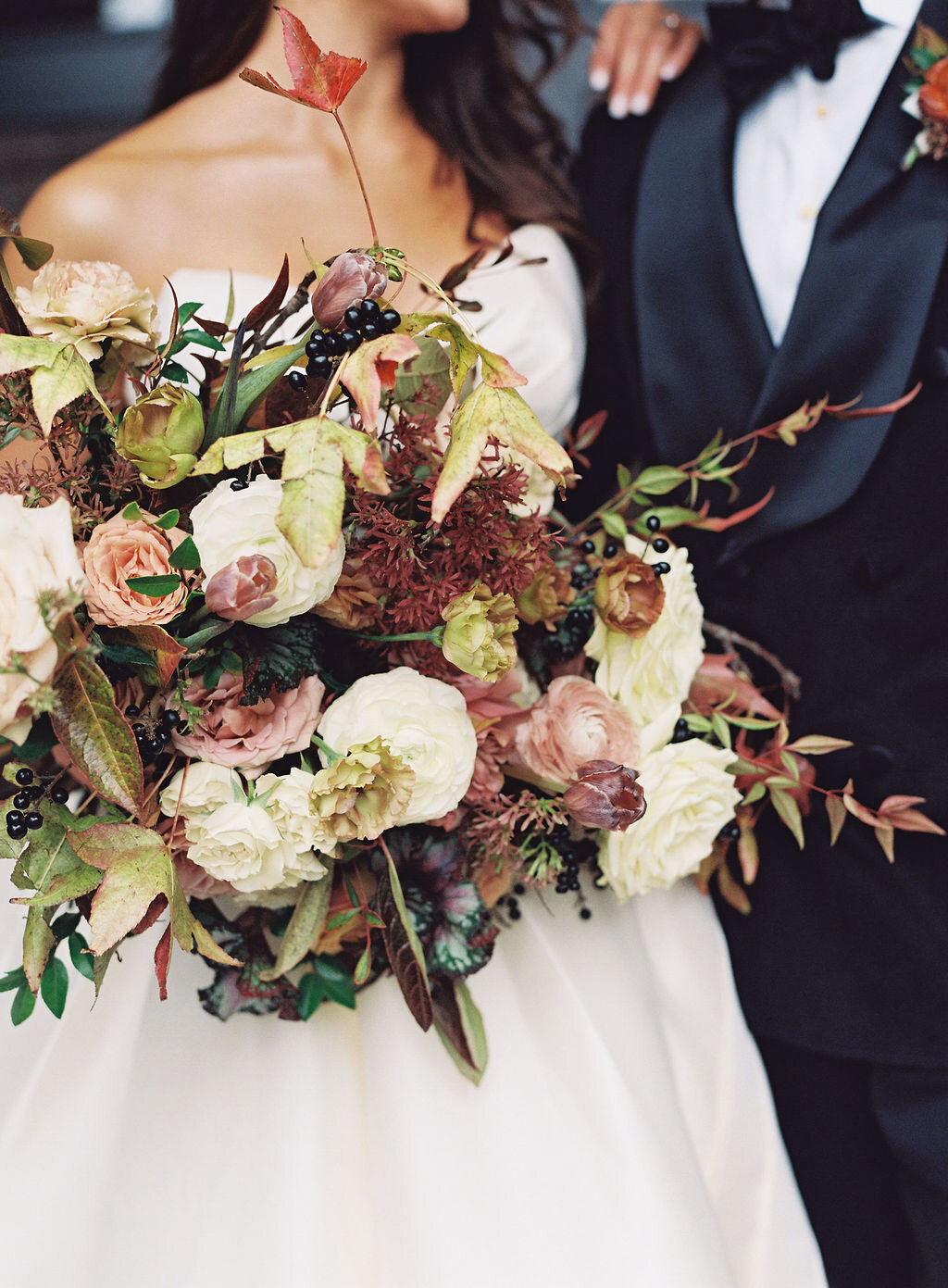 Lush, wild, untamed fall bridal bouquet with a muted autumnal color palette, composed of ligustrum berries, dusty pink ranunculus, tulips, garden roses, fall leaves, begonia leaves, and muted textures. Nashville wedding and corporate event floral de…