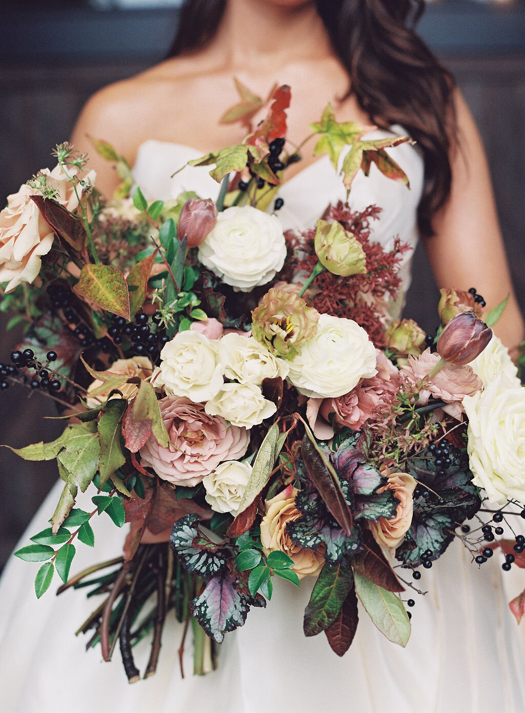 Lush, wild, untamed fall bridal bouquet with a muted autumnal color palette, composed of ligustrum berries, dusty pink ranunculus, tulips, garden roses, fall leaves, begonia leaves, and muted textures. Nashville wedding and corporate event floral de…