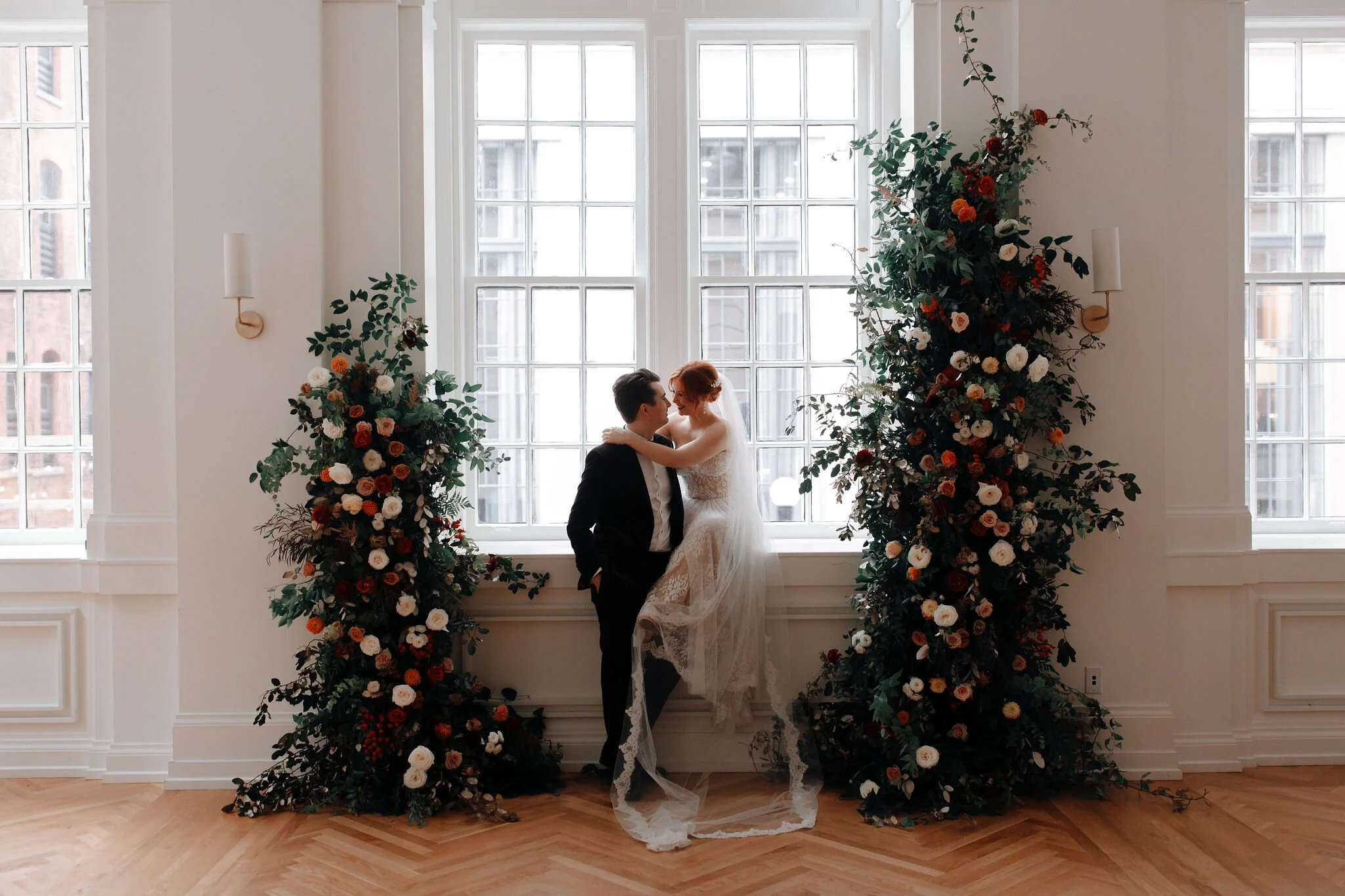 Growing floral installation on either side of the window for the wedding ceremony backdrop, with untamed greenery, rusty orange and burnt sienna garden roses, and lots of texture. Nashville wedding florist at the Noelle hotel.
